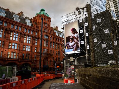 A billboard of the artwork 'Care' by Dola Posh on a busy London road. A black woman sits atop her bed with a toddler sat cross-legged in front of her. She is wearing a floral robe with a burgundy head wrap. The woman combs out her daughter's afro and puts it into twists.