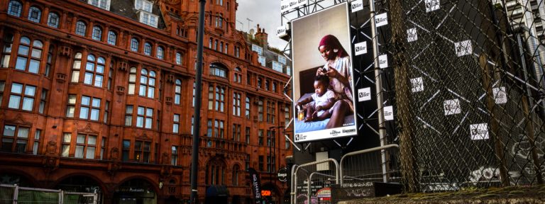 A billboard of the artwork 'Care' by Dola Posh on a busy London road. A black woman sits atop her bed with a toddler sat cross-legged in front of her. She is wearing a floral robe with a burgundy head wrap. The woman combs out her daughter's afro and puts it into twists.