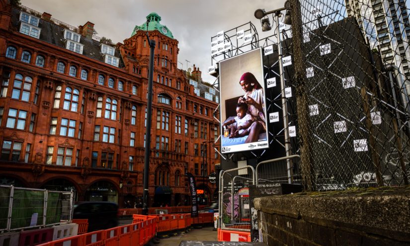 A billboard of the artwork 'Care' by Dola Posh on a busy London road. A black woman sits atop her bed with a toddler sat cross-legged in front of her. She is wearing a floral robe with a burgundy head wrap. The woman combs out her daughter's afro and puts it into twists.