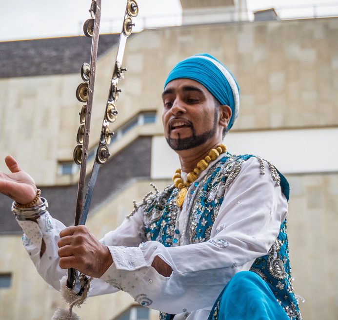 Hardeep Sahota is playing the Chimta with his Bangara music group. He wears a blue headscarf and vest. He is also wearing a white shirt that is adorned with silver beads and sequins. The Chimta is a percussive set of tongs with little bells attached to it.