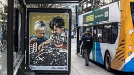 A bus shelter screen of the artwork Last to inherit (2020) by HEYDT in Manchester. A parody of an old children's building blocks advert. Pictured are two smiling white children playing with what appears to be a miniature scale city in ruins. Debris and vehicles are strewn across the play-set.