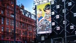 A billboard of the artwork 'Last to inherit' (2020) by HEYDT on a busy London road. A parody of an old children's building blocks advert. Pictured are two smiling white children playing with what appears to be a miniature scale city in ruins. Debris and vehicles are strewn across the play-set.