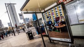 A bus shelter screen of the artwork What We Do (2023) by Becca + Clare in Manchester. A group of people from different backgrounds who vary in age sit in a room together. The room's furniture is constructed from colourful paper and cardboard as if it were the set of a children's show. The cardboard scene details; two children playing with a console game; someone hanging a picture frame; someone knitting and someone lighting candles.