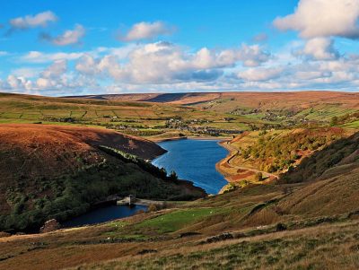A landscape image of Butterley Resevoir. Rolling hills of fields and trees cover the image in warm orange, green and brown tones. There is a winding blue body of water cutting through the middle of the hills. The blue sky above is dotted with white fluffy clouds