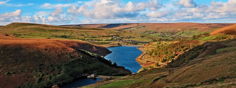 A landscape image of Butterley Resevoir. Rolling hills of fields and trees cover the image in warm orange, green and brown tones. There is a winding blue body of water cutting through the middle of the hills. The blue sky above is dotted with white fluffy clouds