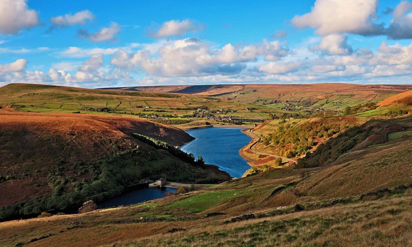 A landscape image of Butterley Resevoir. Rolling hills of fields and trees cover the image in warm orange, green and brown tones. There is a winding blue body of water cutting through the middle of the hills. The blue sky above is dotted with white fluffy clouds