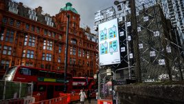 A billboard of the artwork ‘A house upside down’ (2023) by Richard Woods on a busy London road. Four geometric blue houses with grey roofs and orange doors. Three stand upright and one is upside down.