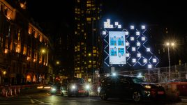 A billboard of the artwork ‘A house upside down’ (2023) by Richard Woods on a busy London road at night. Four geometric blue houses with grey roofs and orange doors. Three stand upright and one is upside down.