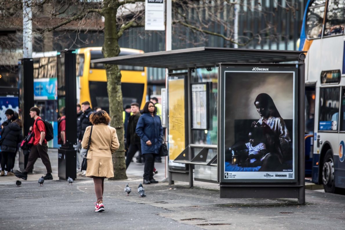 A bus shelter screen of the artwork ‘Care’ (2021) by Dola Posh in Manchester. A black woman sits atop her bed with a toddler sat cross-legged in front of her. She is wearing a floral robe with a burgundy head wrap. The woman combs out her daughter's afro and puts it into twists.