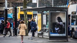 A bus shelter screen of the artwork ‘Care’ (2021) by Dola Posh in Manchester. A black woman sits atop her bed with a toddler sat cross-legged in front of her. She is wearing a floral robe with a burgundy head wrap. The woman combs out her daughter's afro and puts it into twists.