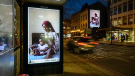 Two billboards of the artwork 'Care' by Dola Posh on a busy London road at night. A black woman sits atop her bed with a toddler sat cross-legged in front of her. She is wearing a floral robe with a burgundy head wrap. The woman combs out her daughter's afro and puts it into twists.