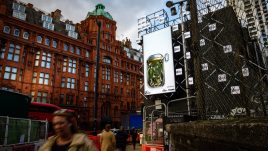 A billboard of the artwork 'Forced into a ‘TickBox’: an Acceptable Method of Gaslighting?' (2023) by Hugh Malyon on a busy London road. A bird’s eye view of what appears to be an open tin of sardines. Stuffed inside are numerous green tinged images of Hugh Maylon (the artist). The many clones of Hugh are uncomfortably crammed together.