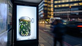 A bus shelter screen of the artwork 'Forced into a ‘TickBox’: an Acceptable Method of Gaslighting?' (2023) by Hugh Malyon on a busy London road. A bird’s eye view of what appears to be an open tin of sardines. Stuffed inside are numerous green tinged images of Hugh Maylon (the artist). The many clones of Hugh are uncomfortably crammed together.