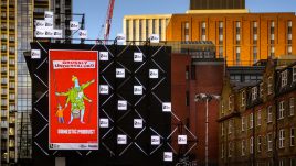 A billboard of the artwork ‘GROSSLY UNDERVALUED DOMESTIC PRODUCT’ (2022) by Bobby Baker on a busy London road. A colourful sketched character on a red background. The central figure is adorned with cooking utensils, washing and other domestic symbols. She has several arms; one holds a bulging shopping basket and another arm leans on a walking cane. A child's hand tightly grasps onto another hand and one arm holds a large flag with text across it. The text reads: 'GROSSLY UNDERVALUED DOMESTIC PRODUCT'.