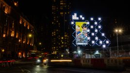 A billboard of the artwork 'Last to inherit' (2020) by HEYDT on a busy London road at night. A parody of an old children's building blocks advert. Pictured are two smiling white children playing with what appears to be a miniature scale city in ruins. Debris and vehicles are strewn across the play-set.