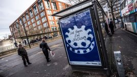 A bus shelter screen of the artwork ‘Let Them Eat Nothing’ (2023) by Sarah Maple in Manchester. A blue and white frosted cake sits against a blue background. The cake is decorated in alternating rosettes of blue and white frosting. Piped onto the cake in cursive lettering are the words: 'Let them Eat Nothing'.