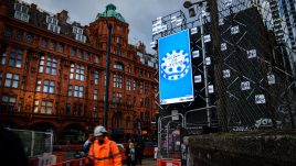 A billboard of the artwork ‘Let Them Eat Nothing’ (2023) by Sarah Maple on a busy London road. A blue and white frosted cake sits against a blue background. The cake is decorated in alternating rosettes of blue and white frosting. Piped onto the cake in cursive lettering are the words: 'Let them Eat Nothing'.