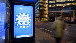 A bus shelter screen of the artwork ‘Let Them Eat Nothing’ (2023) by Sarah Maple on a busy London road at night. A blue and white frosted cake sits against a blue background. The cake is decorated in alternating rosettes of blue and white frosting. Piped onto the cake in cursive lettering are the words: 'Let them Eat Nothing'.