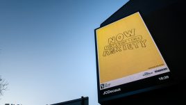 A billboard of the artwork ‘NOW! That’s What I Call Anxiety’ (2022) by Trackie McLeod against the sky at sunset. A vibrant yellow background fills the image. Text in scrawled bold bubble lettering reads: 'NOW THAT'S WHAT I CALL ANXIETY'.