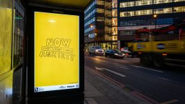 A bus shelter screen of the artwork ‘NOW! That’s What I Call Anxiety’ (2022) by Trackie McLeod on a busy London road at night. A vibrant yellow background fills the image. Text in scrawled bold bubble lettering reads: 'NOW THAT'S WHAT I CALL ANXIETY'.