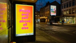 A bus shelter screen of the artwork ‘Profit and Self’ (2022) by S Mark Gubb on a busy London road at night. The background is a gradient of yellow to orange. In large fuchsia letters, the text reads: 'Profit and self above all else'.