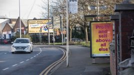 A bus shelter screen of the artwork ‘Profit and Self’ (2022) by S Mark Gubb in Manchester. The background is a gradient of yellow to orange. In large fuchsia letters, the text reads: 'Profit and self above all else'.