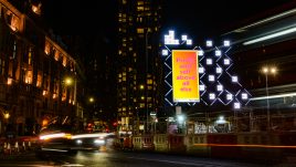 A billboard of the artwork ‘Profit and Self’ (2022) by S Mark Gubb on a busy London road at night. The background is a gradient of yellow to orange. In large fuchsia letters, the text reads: 'Profit and self above all else'.