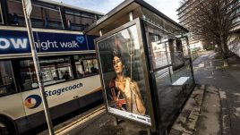 A bus shelter screen of the artwork ‘Rue The Waves’ (2022) by Natasha Klutch in Manchester. A painted portrait of a regal looking woman in patriotic British colours. She is holding a bent trident, a round Union Jack shield and a helmet with a lion on it. She has a black eye and is covered in tomato juice.