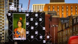 A billboard of the artwork ‘Rue The Waves’ (2022) by Natasha Klutch on a busy London road. A painted portrait of a regal looking woman in patriotic British colours. She is holding a bent trident, a round Union Jack shield and a helmet with a lion on it. She has a black eye and is covered in tomato juice.