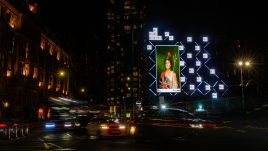 A billboard of the artwork ‘Rue The Waves’ (2022) by Natasha Klutch on a busy London road at night. A painted portrait of a regal looking woman in patriotic British colours. She is holding a bent trident, a round Union Jack shield and a helmet with a lion on it. She has a black eye and is covered in tomato juice.