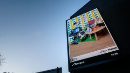 A billboard of the artwork ‘What We Do’ (2023) by Becca + Clare against a sunset. A group of people from different backgrounds who vary in age sit in a room together. The room's furniture is constructed from colourful paper and cardboard as if it were the set of a children's show. The cardboard scene details; two children playing with a console game; someone hanging a picture frame; someone knitting and someone lighting candles.