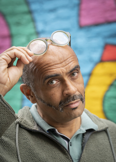 Martin Glover looks into the camera and lifts his glasses to rest them on his head. He is standing in front of a colourful abstract wall.