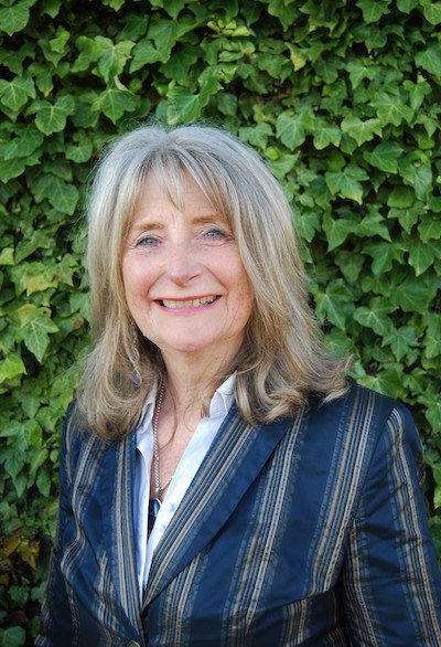 Angela Sandwith wearing a navy blazer and white shirt standing in front of a hedge. She is smiling and her hair is shoulder length