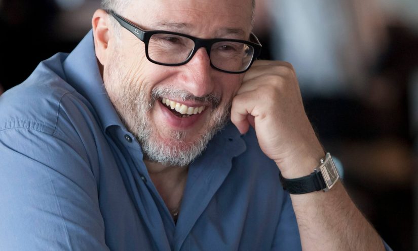 Tony Heaton wearing a pale blue shirt. His leans his face into his left hand and smiles into the camera.