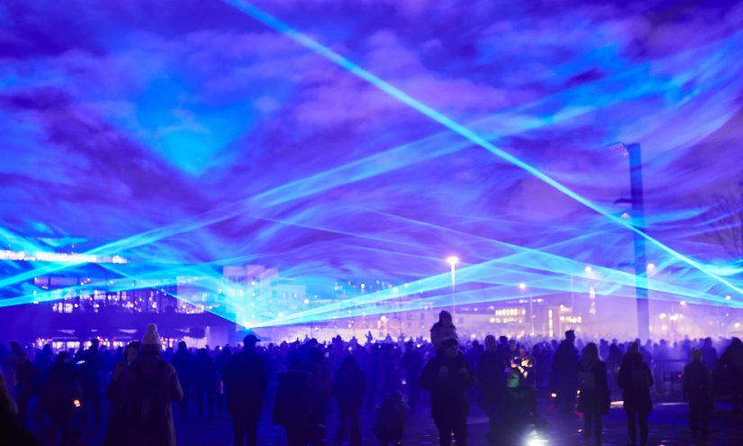 A blue light projection artwork fills the sky above a crowd of people.