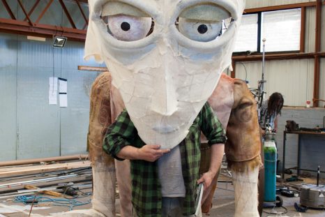 An image of a man in a green and black checkered shirt. The large torso of a puppet made from chicken wire and stuffing can be seen behind him. He is holding a huge partially finished puppet head infront of his face. The wire skelleton of the human head is mostly covered in white cloth. It has eyeballs and eyelids installed aswell.