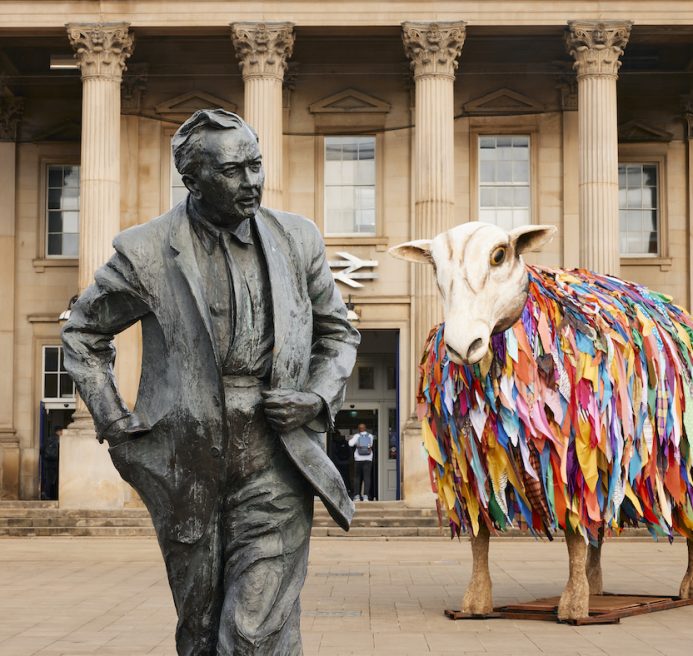 Aina, the Mother Sheep Sculpture, in front of St George’s Square. The statue of Harold Wilson is in the foreground and the station is in the background.