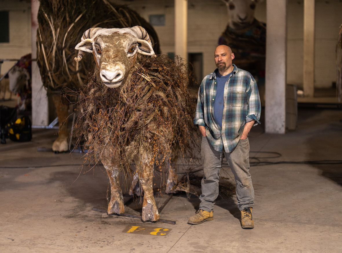 Dave standing with one of the singing sheep sculptures in the HERD workshop. He is wearing a blue checkered shirt and has his hands in his pockets. The sheep is to his left.