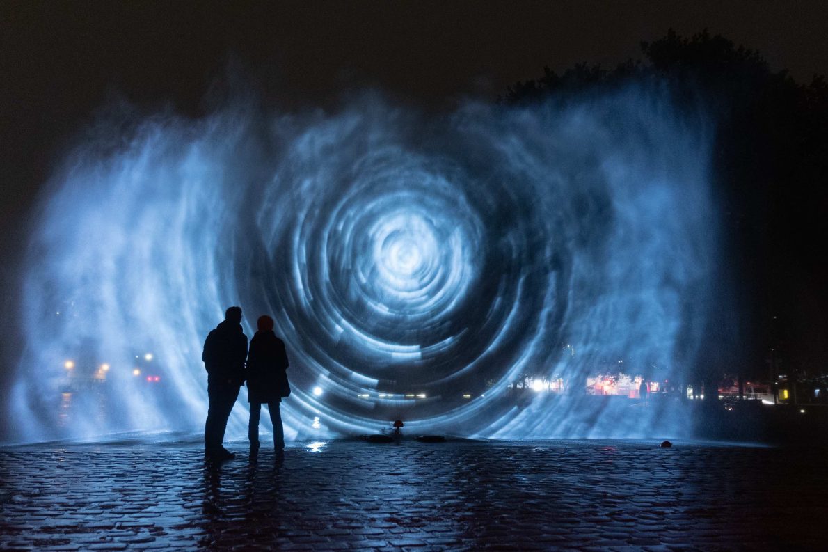 Two people stand in front of a blue projection. It is abstract, but looks somewhat like a black hole. It is night time and the street is cobbled.