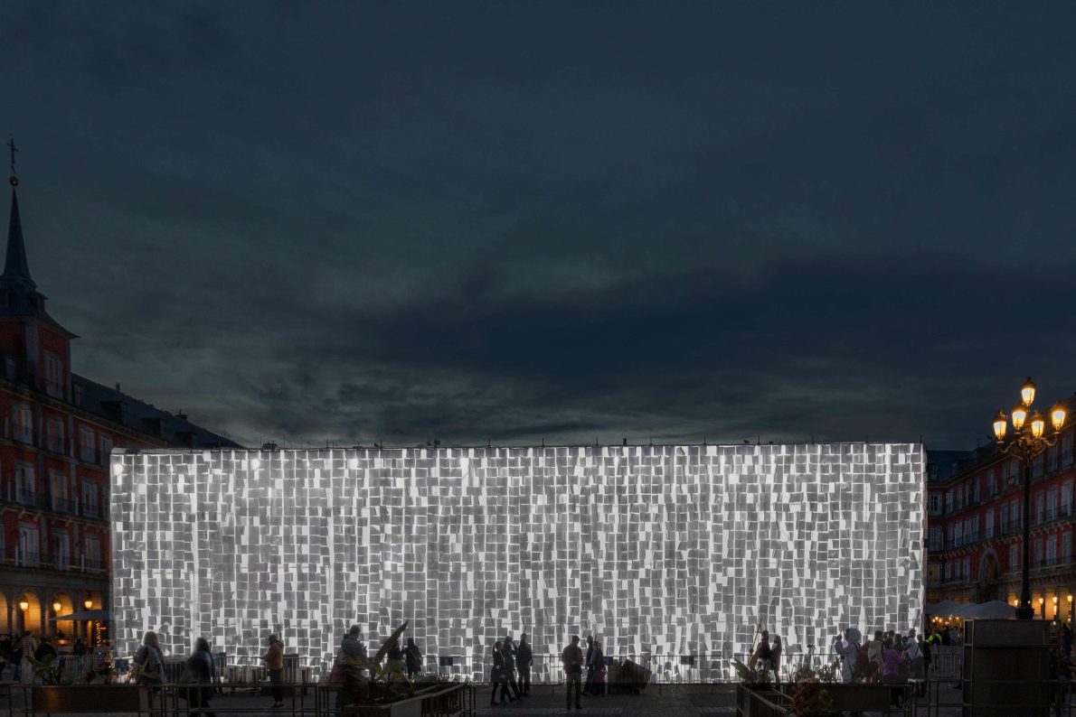 A large-scale illuminated wall covered in thousands of notebooks. You can see the silhouettes of people standing beneath the art installation, writing things in the blank pages. It is dusk.