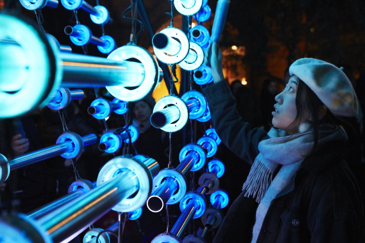A woman touches the art installation. She is seen from the side wearing a beret and a scarf. The artwork has a metal frame and lots of hoops attached to it. The hoops are illuminated and glow blue.