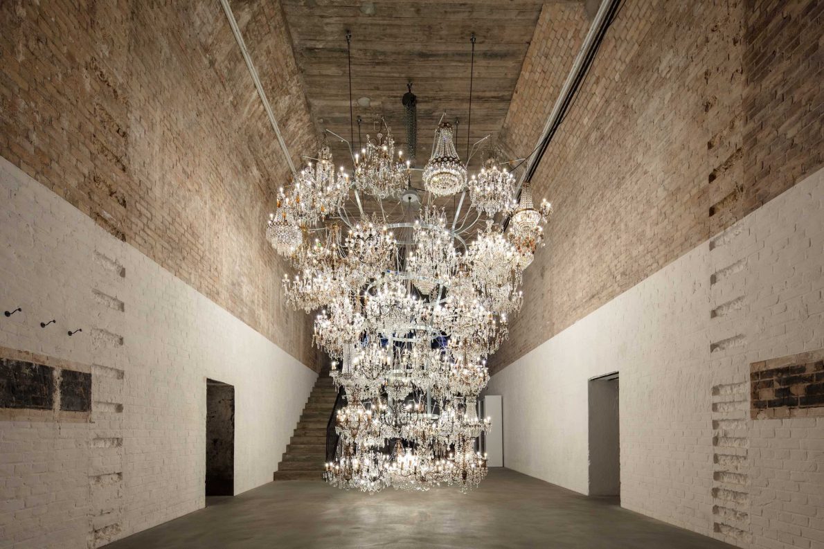 Large-scale chandelier in a room with brick walls and high ceilings. The chandelier is made up of multiple other chandeliers and it is lit up, illuminating the room.