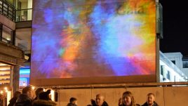 The artwork 'Holi' by OCUBO. Outside the Gala Theatre in Durham at night. A projection of colourful powders splash across the wall, reacting to the movements of the crowd below.
