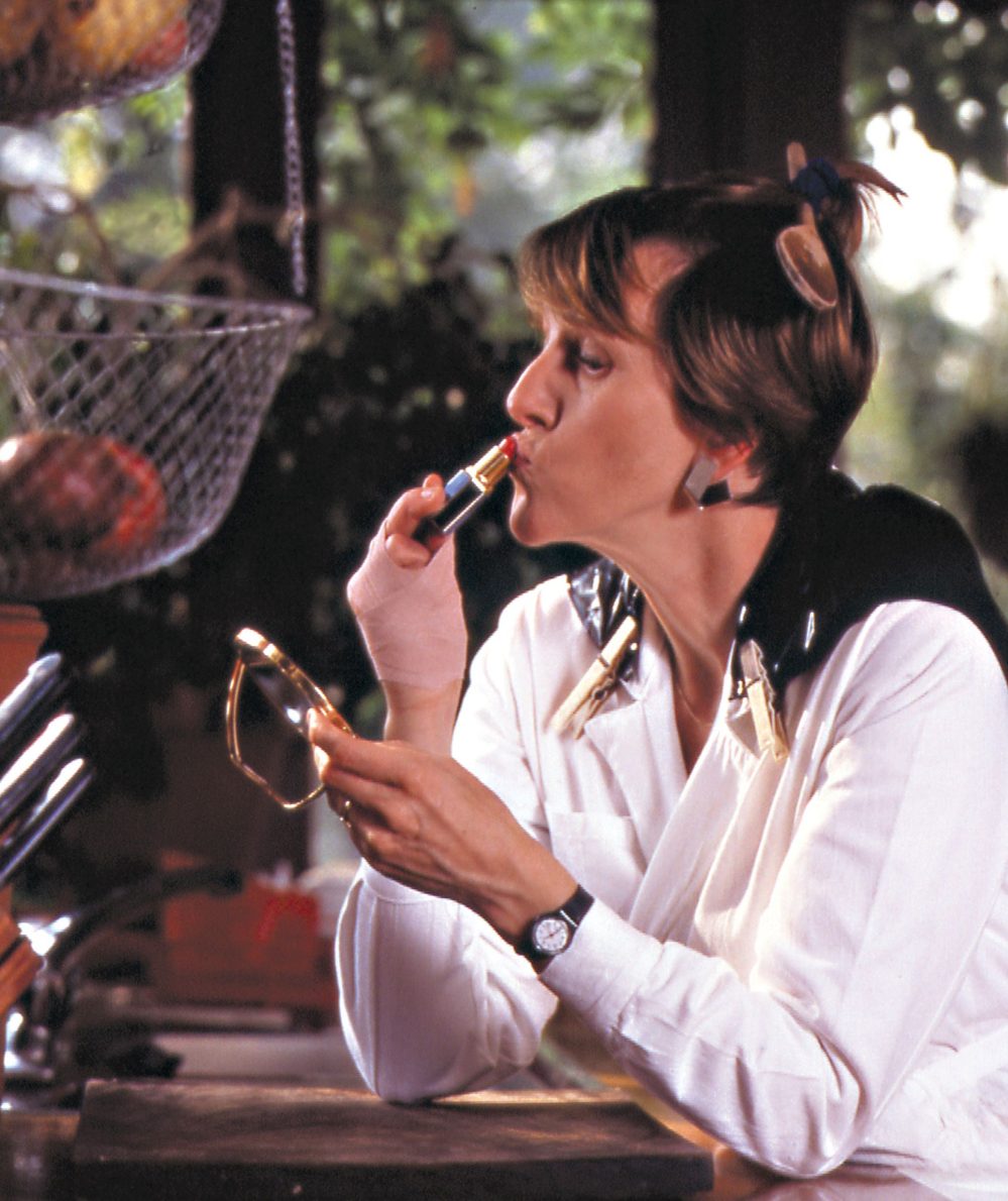 Bobby Baker is photographed applicing red lipstick in a kitchen with kitchen utensils in her hair.