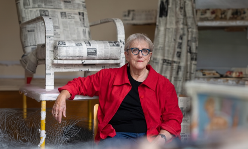 A photograph of bobby Baker infront of one of her aartworks. She is wearing a red shirt and blue glasses