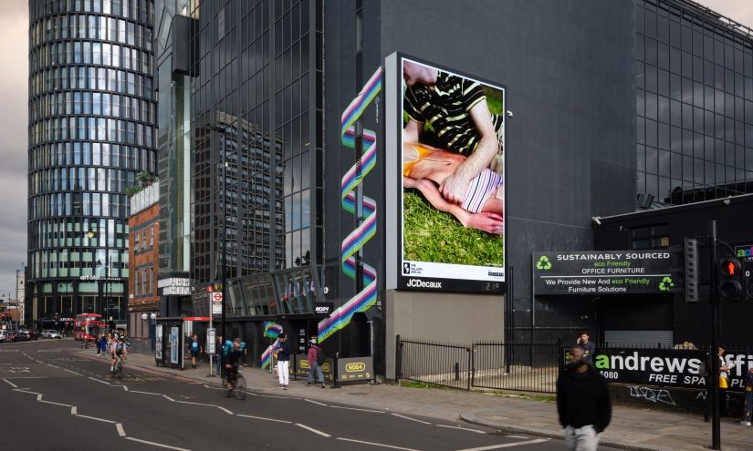 A city street with a large digital billboard displaying artwork of two people lying on grass.