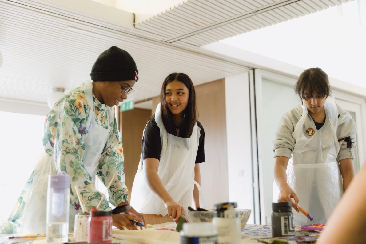 Three young people wearing aprons paint together at a table, smiling and focused on their artwork in a bright indoor space.
