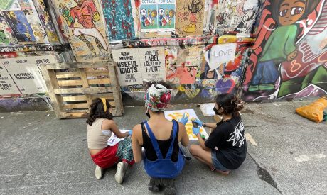 Three young individuals sit on the ground in front of a colorful graffiti-covered wall, engaging in a creative activity involving spray painting