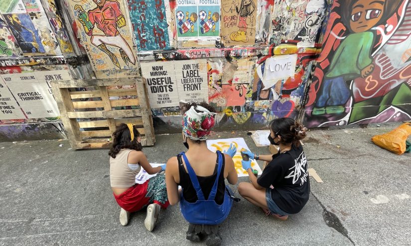 Spray painting with Tasnim Mahdy. School 2024. Photo by Beth King Three young individuals sit on the ground in front of a colorful graffiti-covered wall, engaging in a creative activity involving spray painting
