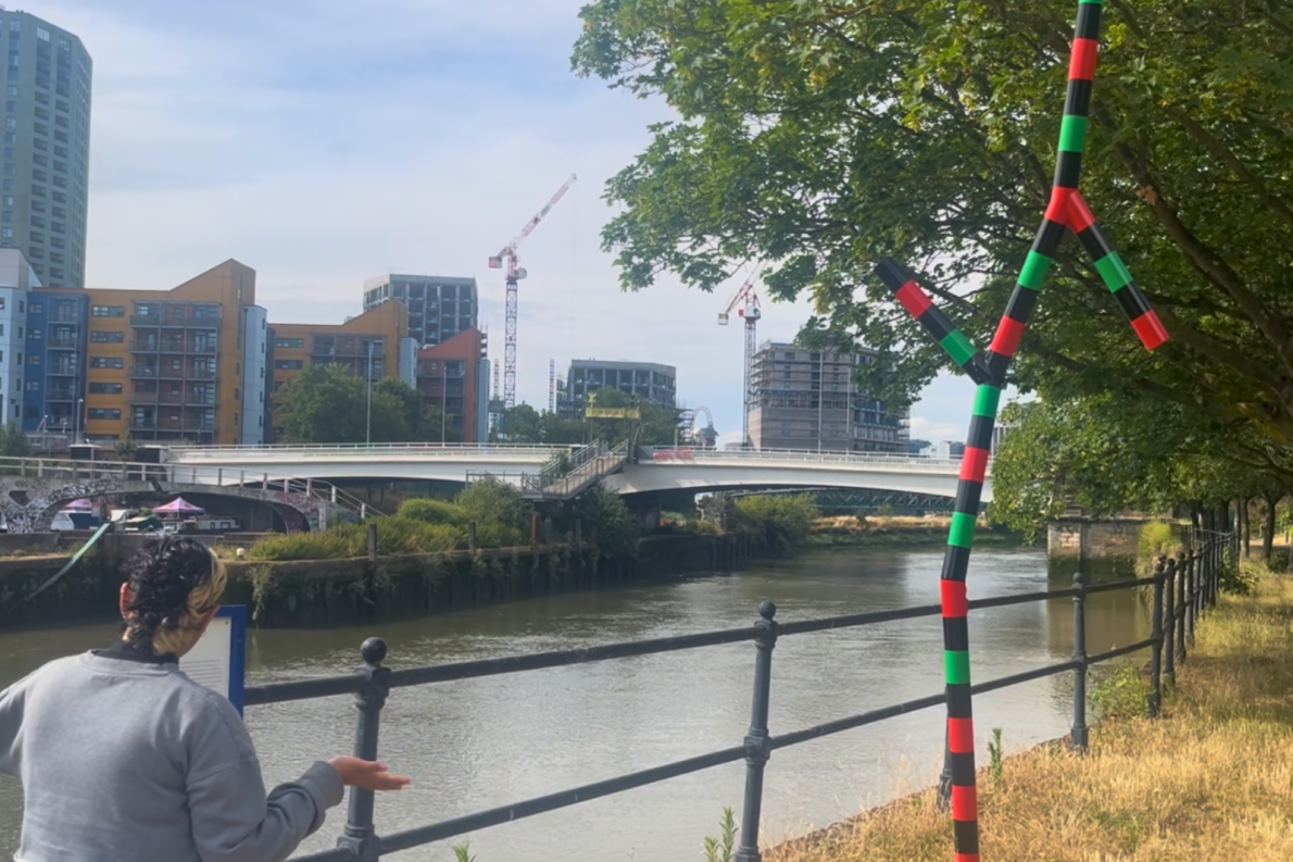 A person observes a riverside with construction cranes, colorful buildings, and a curved bridge. A striped pole sculpture and leafy trees add vibrancy.
